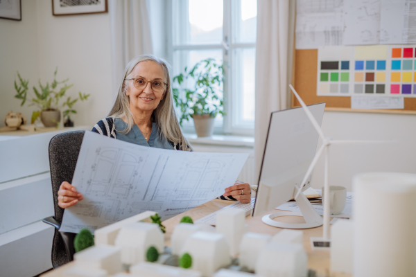 A senior woman eco architect with blueprints working on lapotp in office.