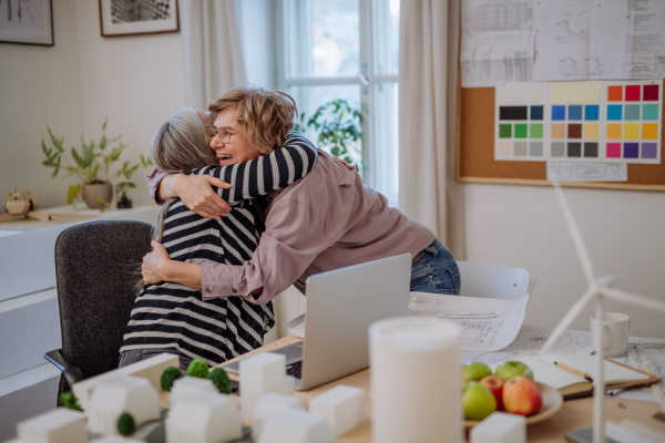 A senior women eco architects with blueprints working on laptop together in office, hugging and having success.