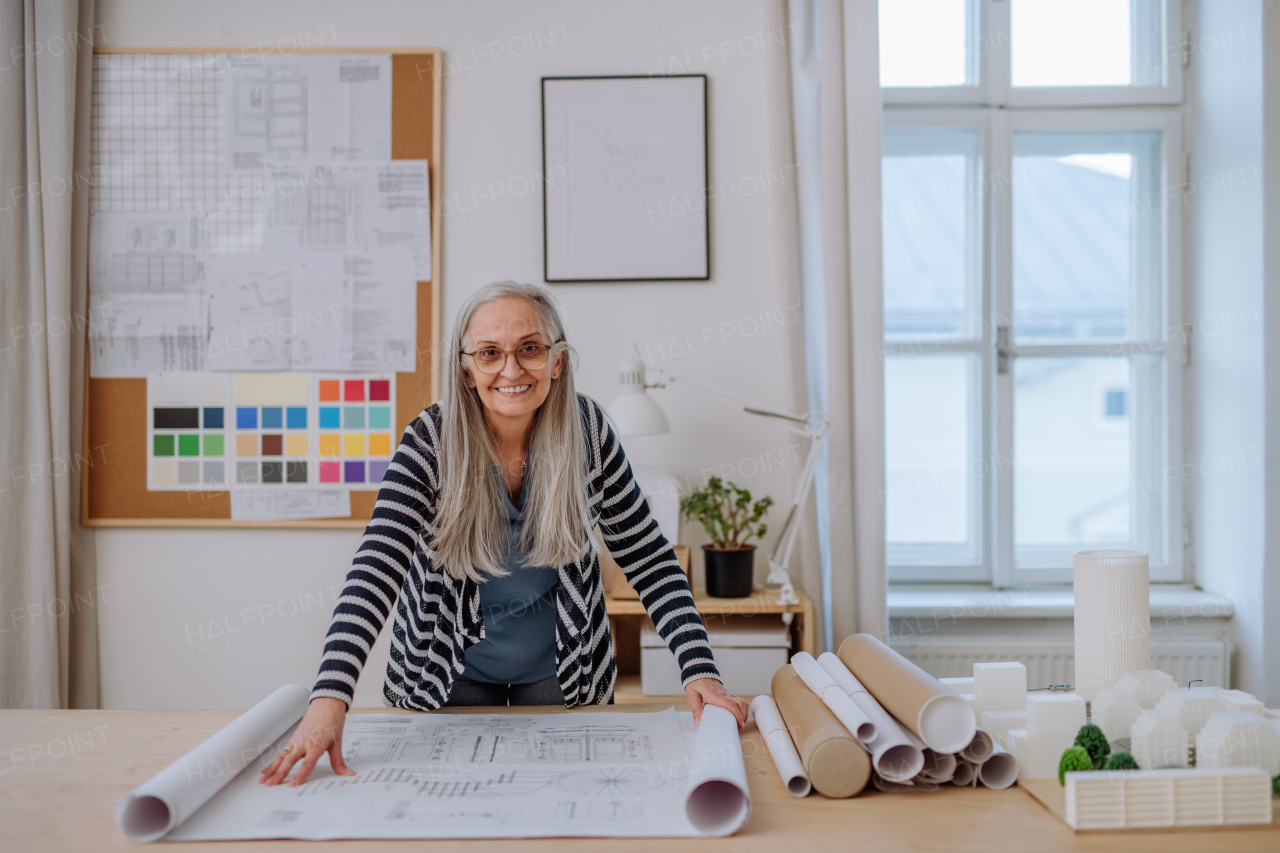 Happy senior woman architect with model of houses standing in office and looking at camera.