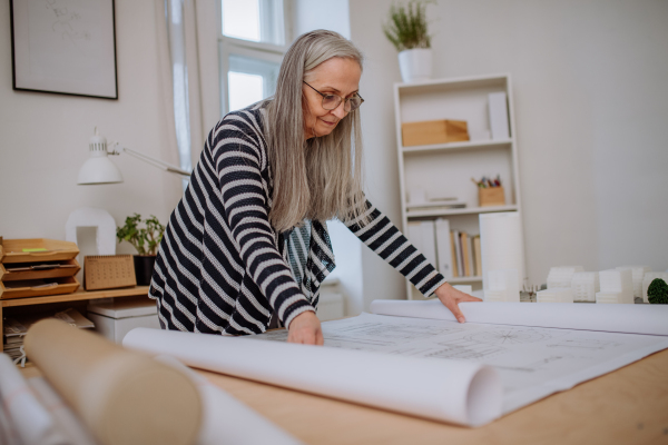 A senior woman architect with model of houses looking at blueprints in office.