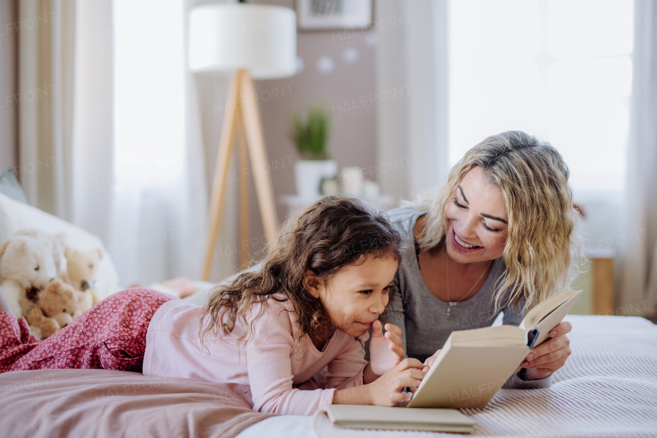 A happy mother with her little daughter lying on bed and reading book at home.