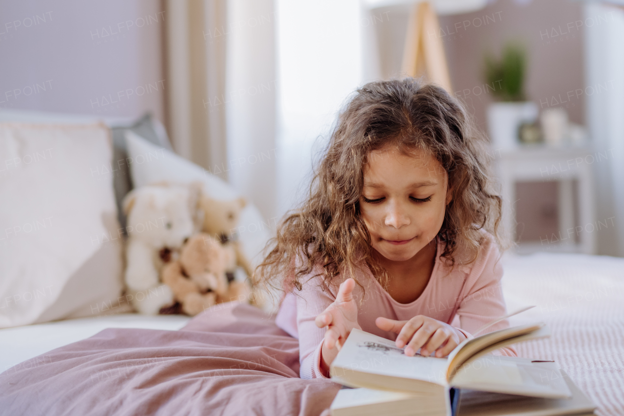 A little girl lying on bed and reading book at home.