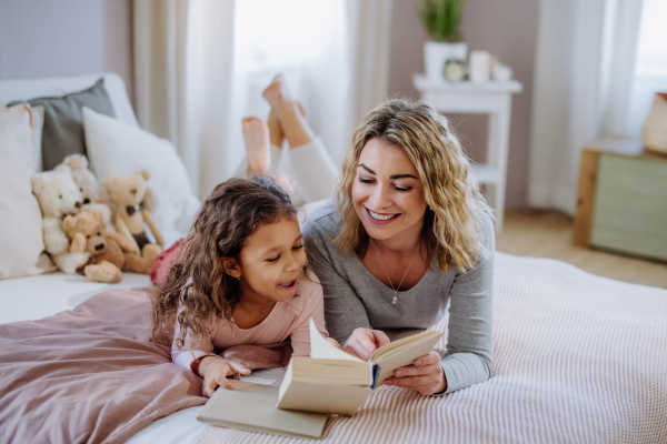 A happy mother with her little daughter lying on bed and reading book at home.