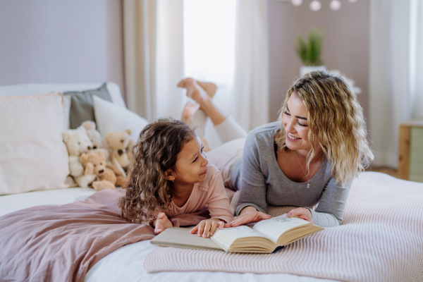 A happy mother with her little daughter lying on bed and reading book at home.