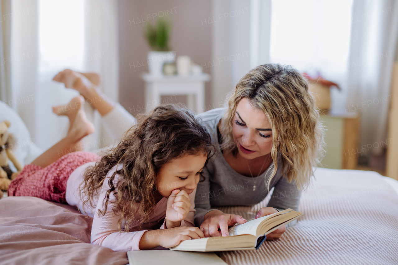 A happy mother with her little daughter lying on bed and reading book at home.