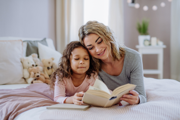 A happy mother with her little daughter lying on bed and reading book at home.