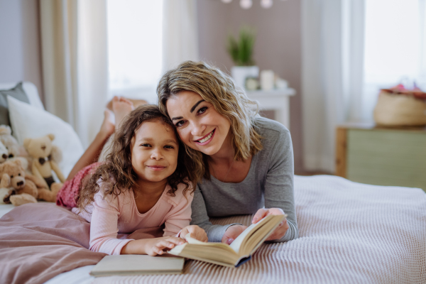 A happy mother with her little daughter lying on bed and reading book at home.