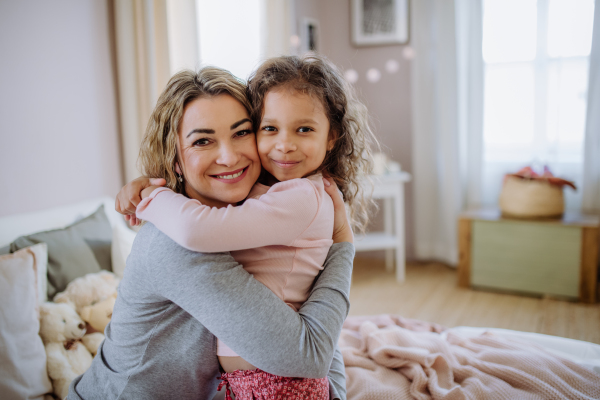 A happy mother with her little daughter hugging together on bed at home and looking at camera.