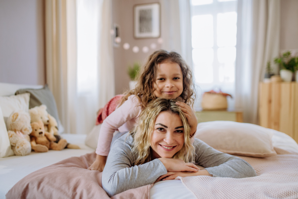 A happy little girl lying on mothers back on bed and looking at camera at home.