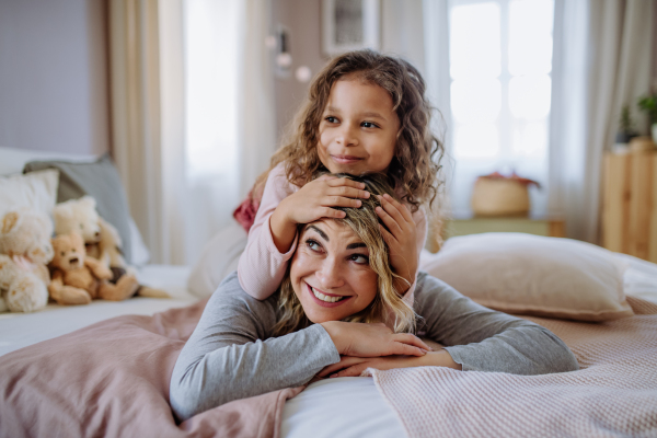 A happy little girl lying on mothers back on bed and looking at camera at home.