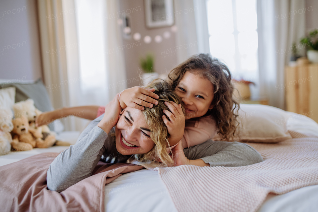 A happy little girl lying on mother's back on bed at home, and looking at camera.