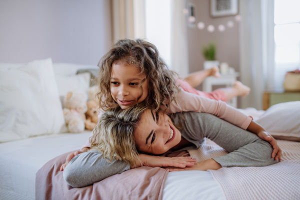 A happy little girl lying on mothers back on bed and looking at camera at home.