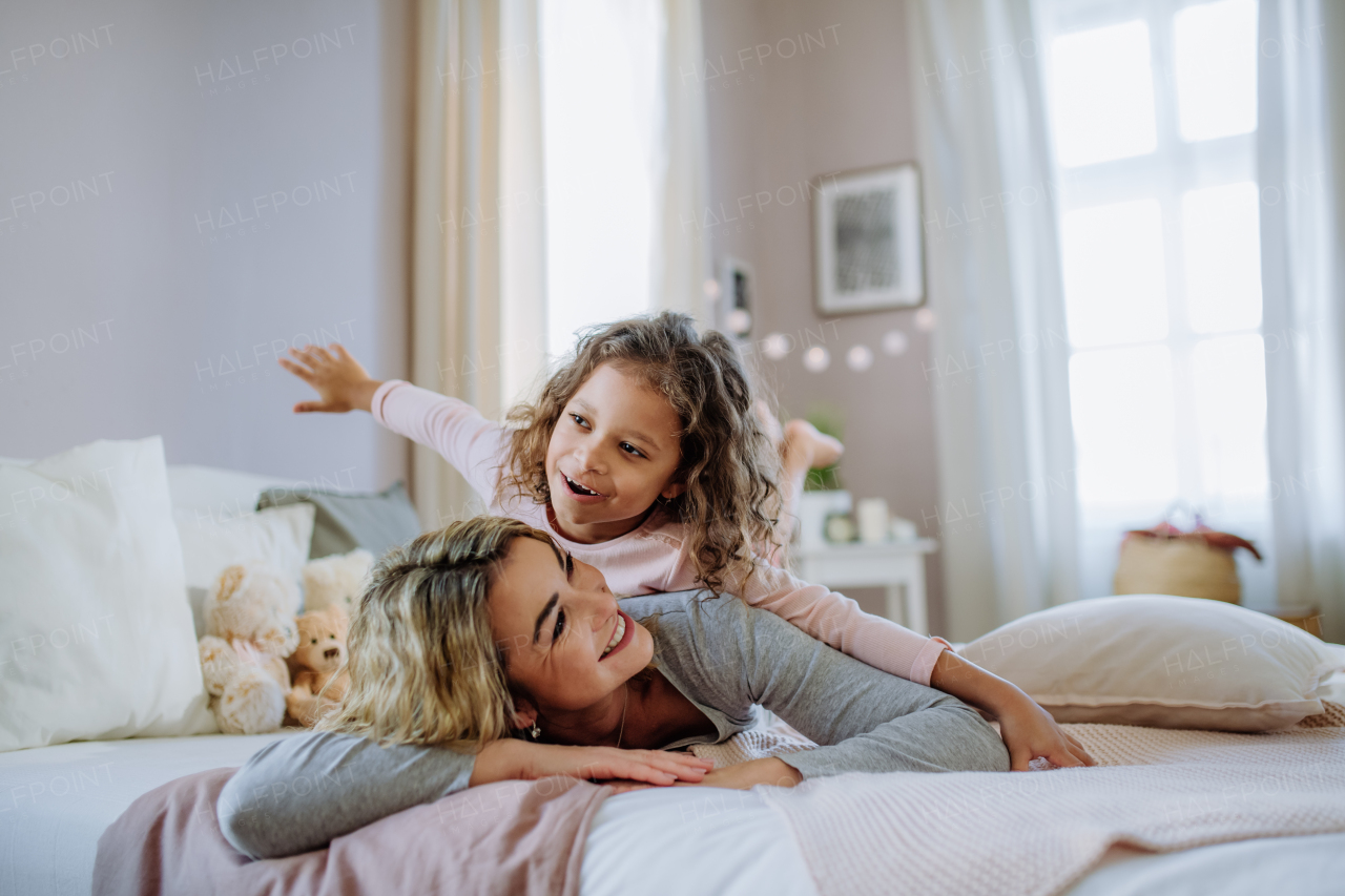 A happy little girl lying on mothers back on bed and looking at camera at home.