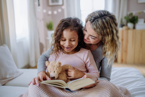 A happy mother with her little daughter sitting on bed and reading book at home.