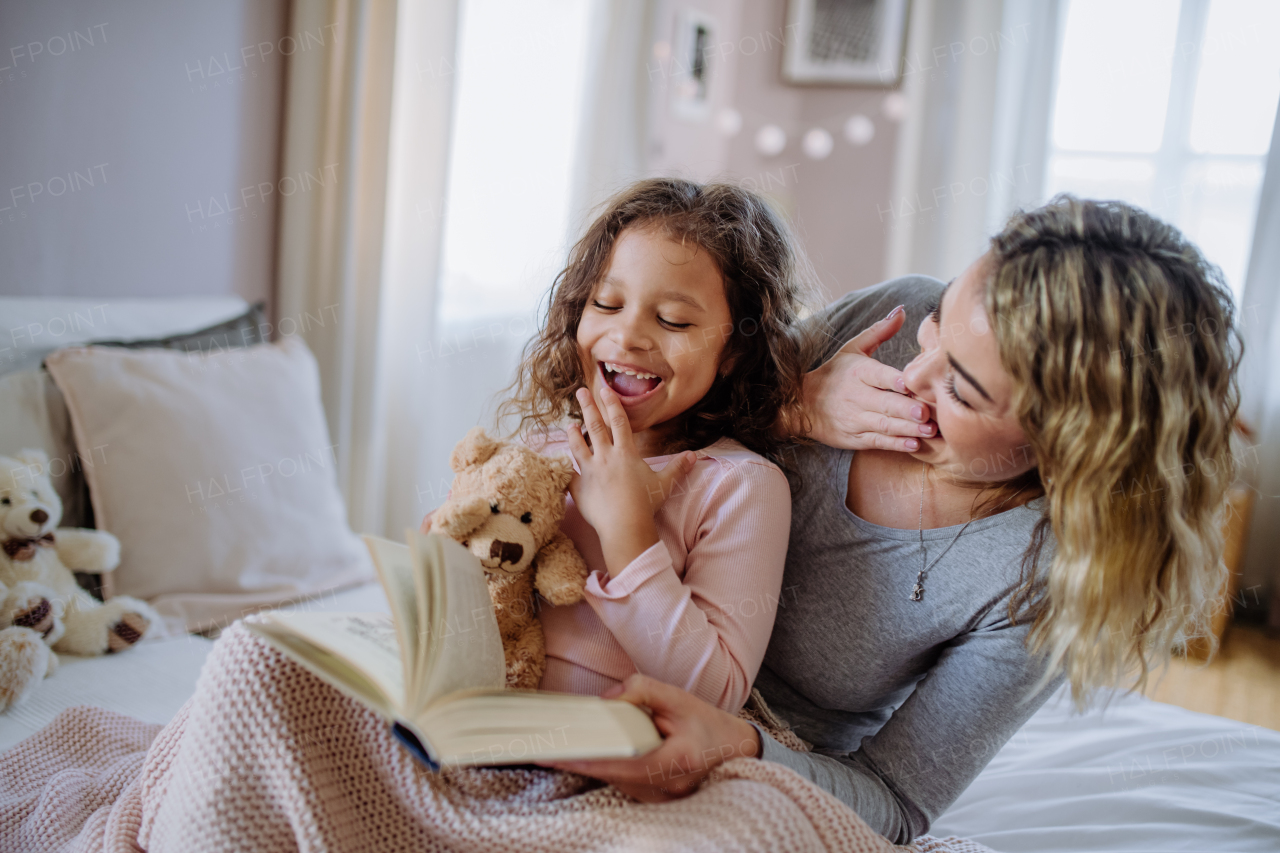 A happy mother with her little daughter sitting on bed and reading book at home.