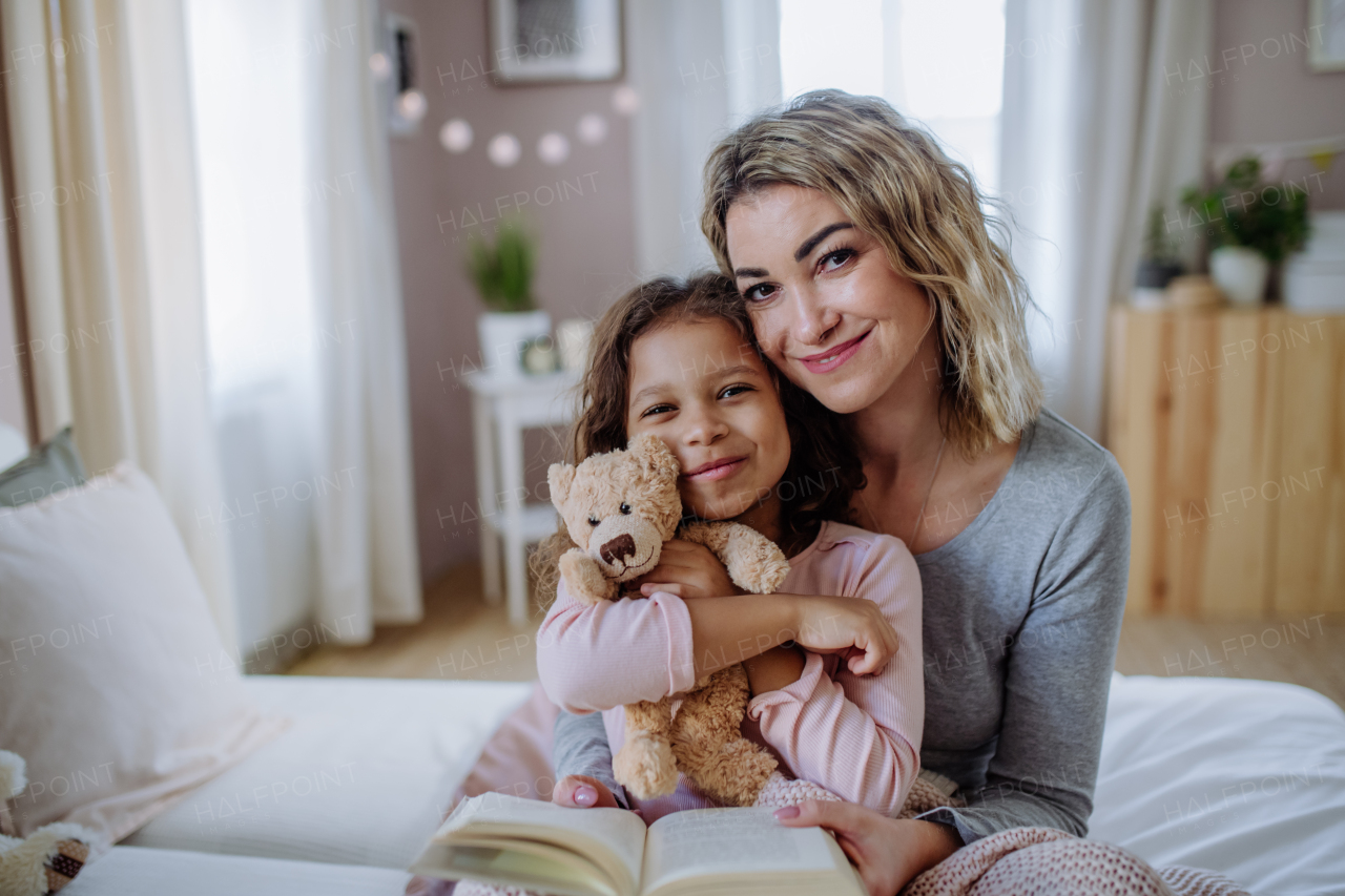 A happy mother with her little daughter hugging together on bed at home and looking at camera.