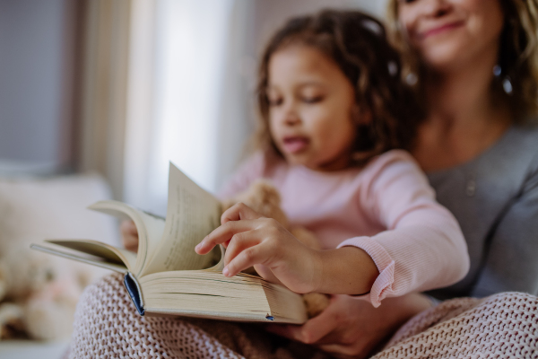 A happy mother with her little daughter sitting on bed and reading book at home.