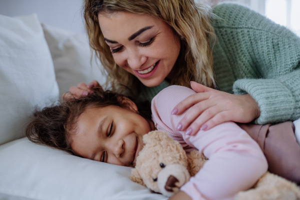 A happy mother stroking her little daughter when lying on bed with her at home.