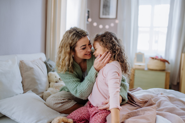 A happy mother stroking her little daughter and enjozing time together on bed at home.