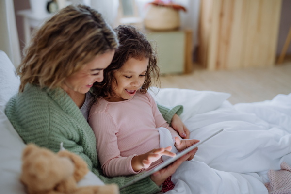 A happy mother with her little daughter lying on bed and using tablet at home.