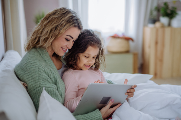 A happy mother with her little daughter lying in bed and using tablet at home.