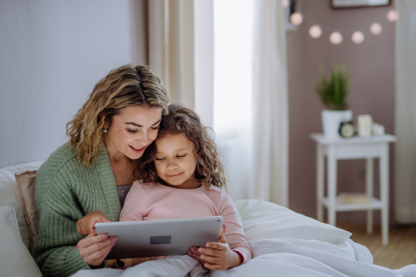 A happy mother with her little daughter lying on bed and using tablet at home.