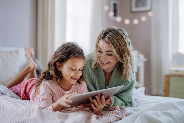 A happy mother with her little daughter lying on bed and using tablet at home.