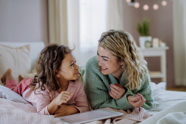 A happy mother with her little daughter lying on bed and using tablet at home.