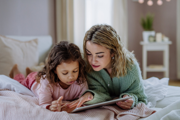 A happy mother with her little daughter lying on bed and using tablet at home.