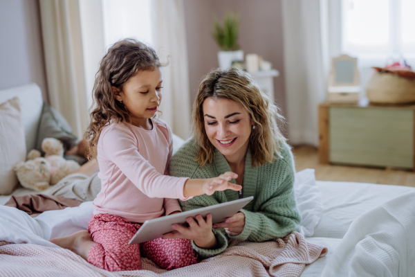 A happy mother with her little daughter lying on bed and using tablet at home.