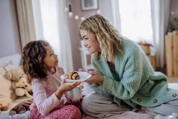 A happy mother with her little daughter having breakfast together in bed at home.