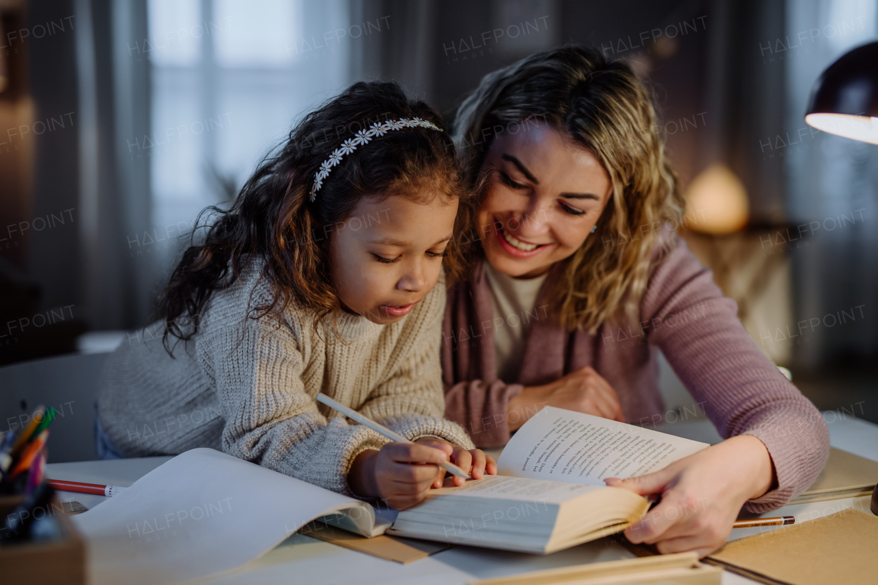 A little multiracial girl doing homework with her mother in evening at home.