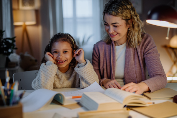 A happy little girl doing homework with her mother and looking at camera in evening at home.