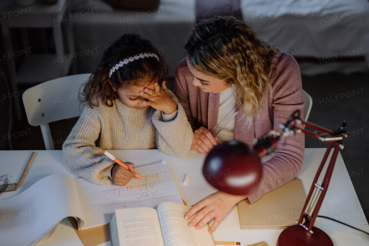 A high angle view of little girl doing homework with her mother in evening at home.