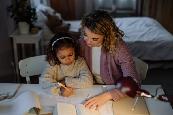 A high angle view of little girl doing homework with her mother in evening at home.