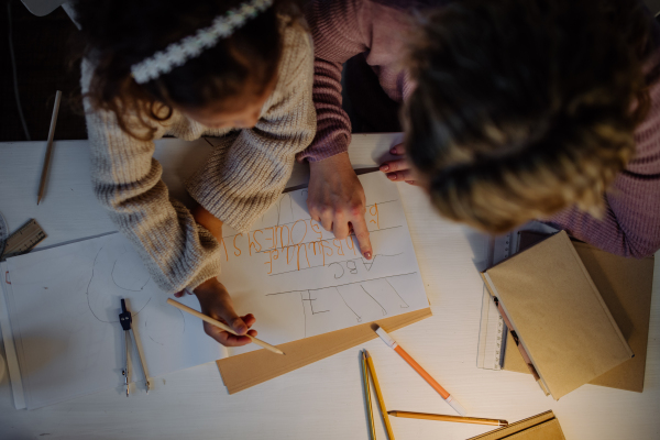 A top view of little girl doing homework with her mother in evening at home.