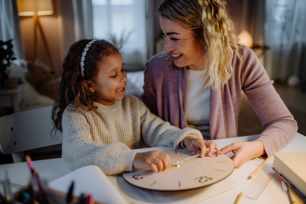 A happy little girl learning time with wooden clock with her mother in evening at home.