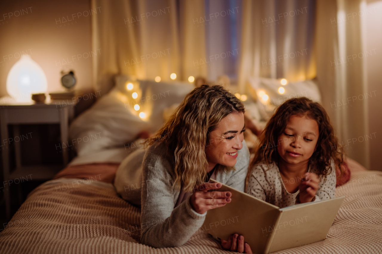 A happy mother with her little daughter lying on bed and reading book in evening at home.