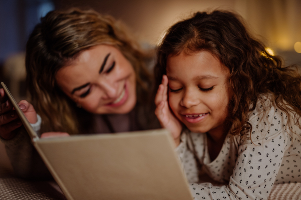 A happy mother with her little daughter lying on bed and reading book in evening at home.