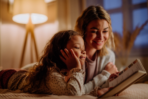 A happy mother with her little daughter lying on bed and reading book in evening at home.