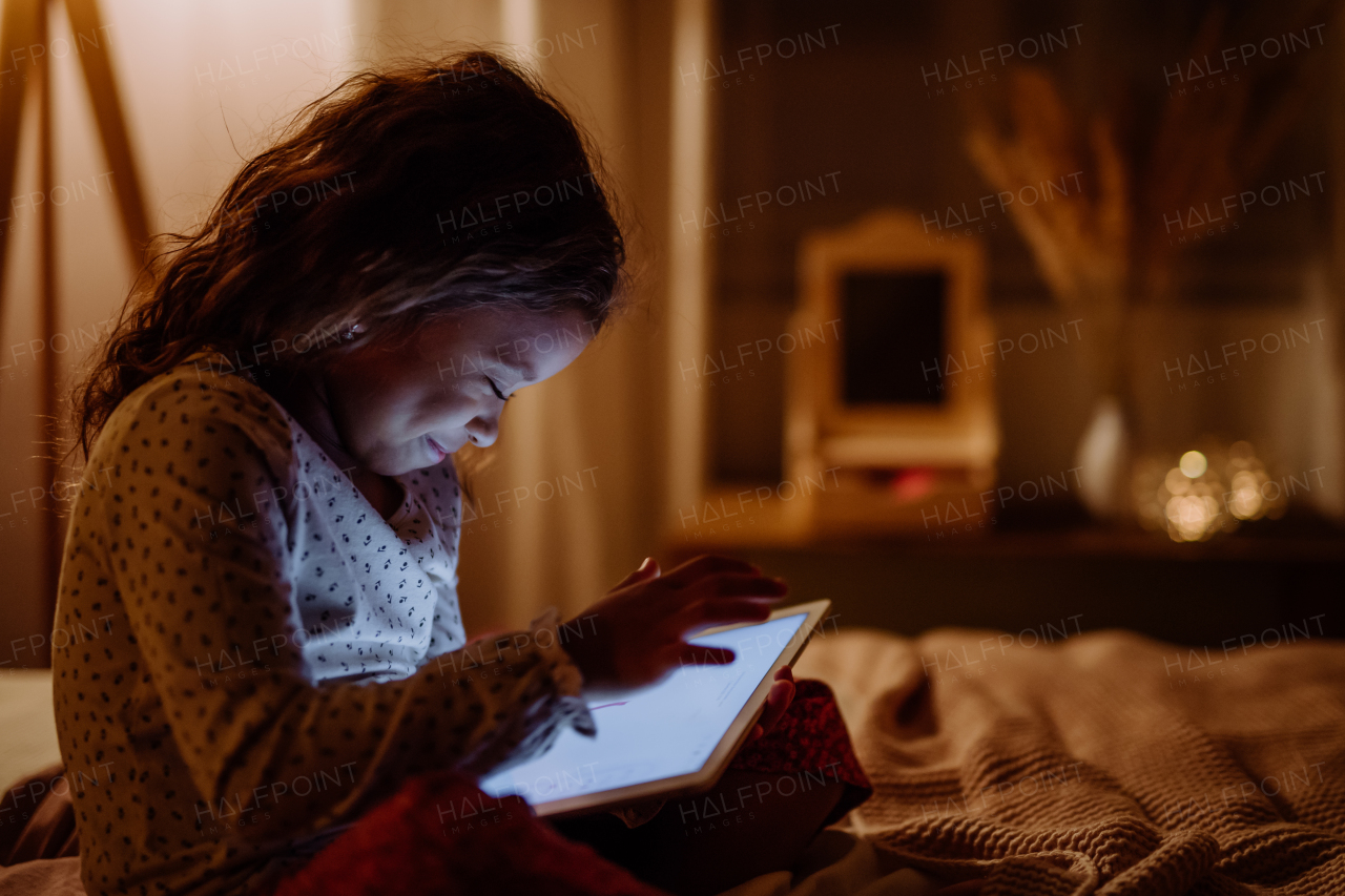 A happy little multiracial girl using tablet on bed in evening at home.