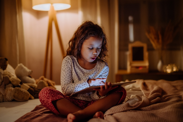 A happy little multiracial girl using tablet on bed in evening at home.