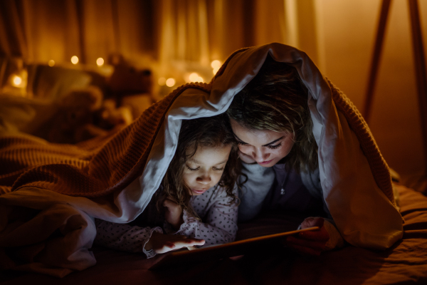 A happy mother with her little daughter lying under blanket and watching movie on tablet at home.