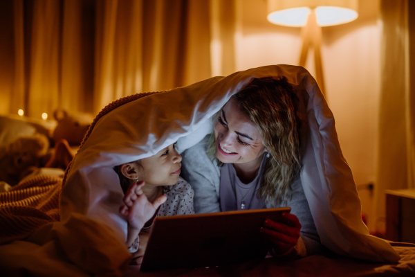 A happy mother with her little daughter lying under blanket and watching movie on tablet at home.