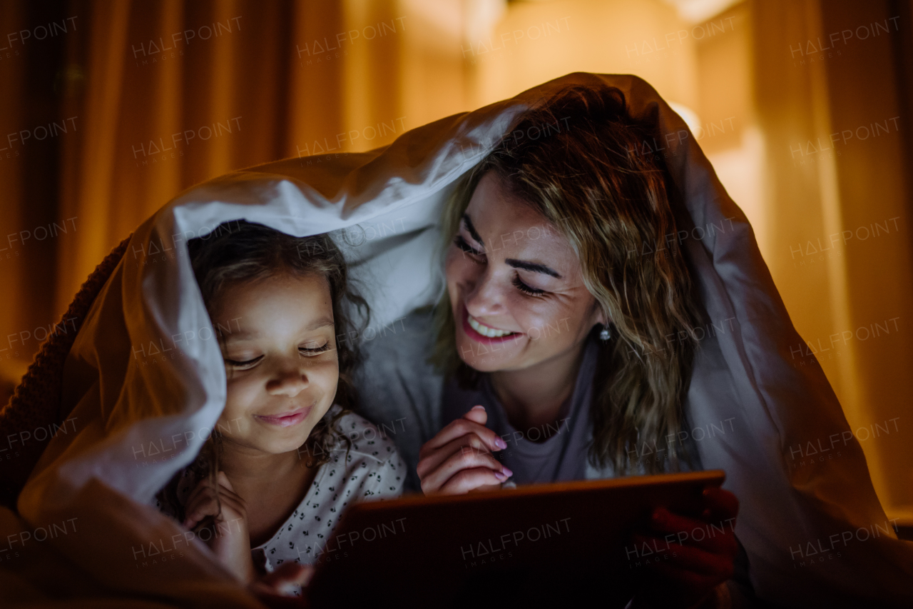 A happy mother with her little daughter lying under blanket and watching movie on tablet at home.