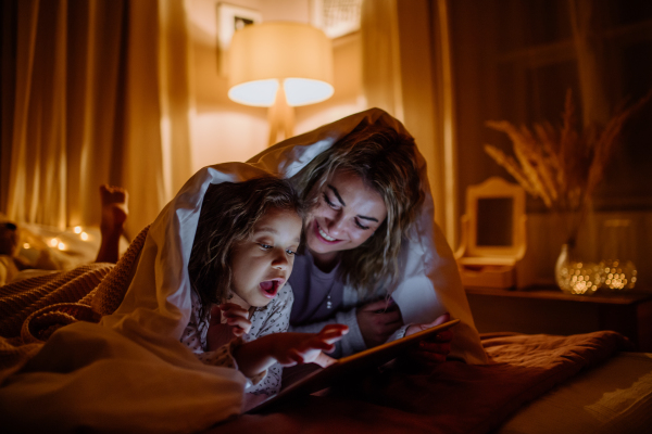 A happy mother with her little daughter lying under blanket and watching movie on tablet at home.