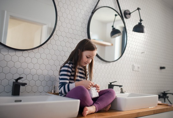 A little girl sitting in bathroom, holding basket and looking for hair clips at home.