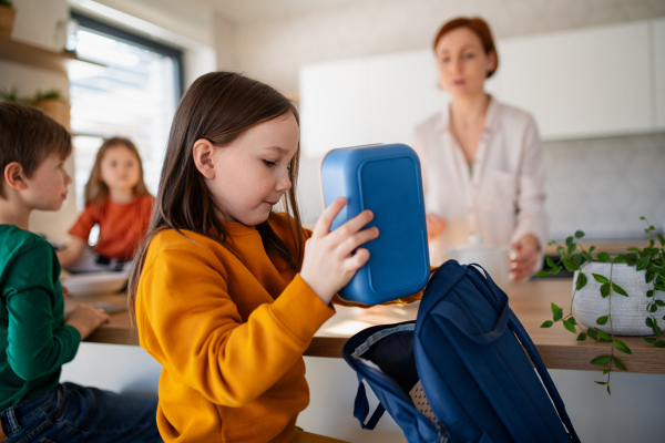 A little girl packing lunch box to backpack in kitchen at home.