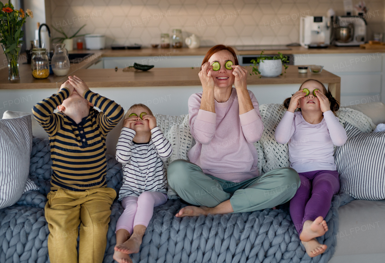 Three little children with a mother sitting on sofa and putting cucumber on faces at home.