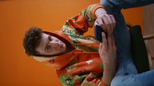 A fashion studio shootingt of a young man in hoodie with smartphone posing over orange background.